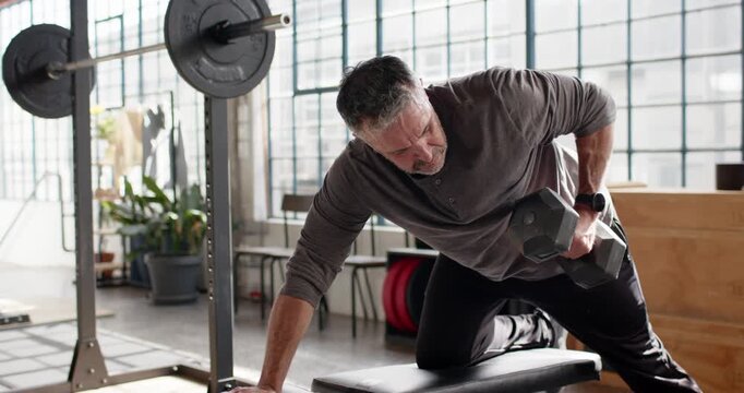 Middle-aged man bracing left hand while kneeling in bright gym, rowing hex dumbbell for strength