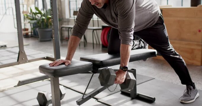 Middle-aged man bracing knee on bench reaching hex dumbbell rowing to ribcage at gym for strength