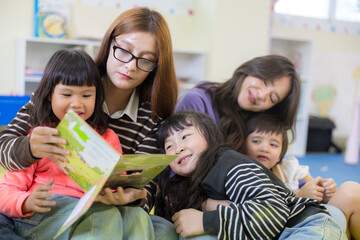 Fototapeta premium An Asian preschool teacher wearing glasses reads a colorful picture book to a group of happy, engaged young children sitting together in a bright daycare classroom.