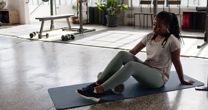African woman post-workout rolling calves with foam roller in leggings on blue mat, copy space