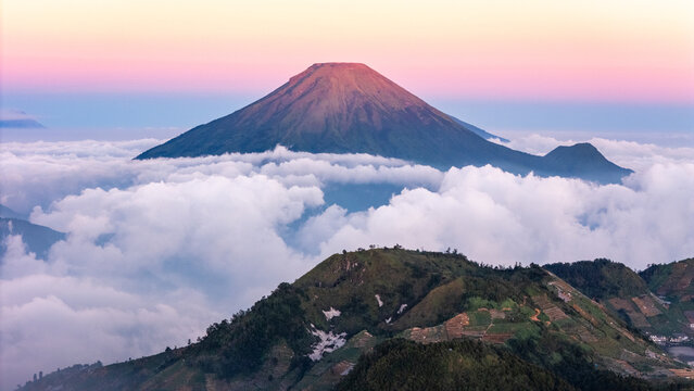 Aerial view of Mount Sindoro rising above a thick sea of clouds with green hills in the foreground under a colorful sunset sky in Indonesia.