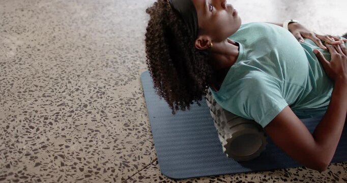 Mid-adult African woman placing foam roller under upper back on mat in home studio, seeking relief