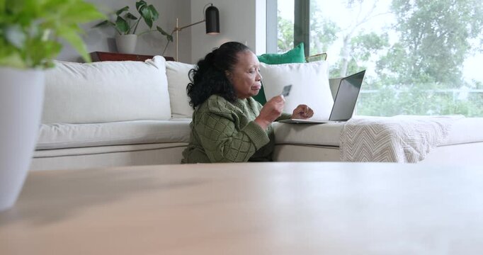 Senior African American woman typing on laptop at home, comparing card for payment and smiling