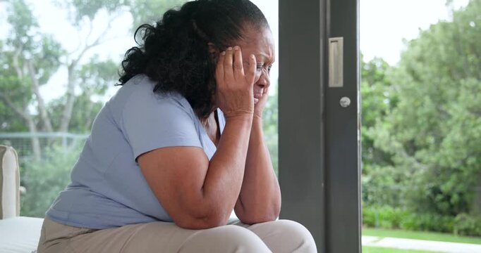 Senior African female on sofa after pressing temples, turning head gazing out glass door at garden