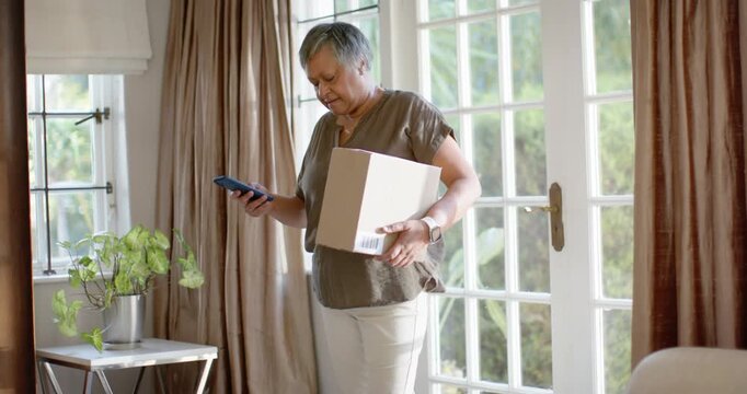 Mature woman holding box scanning code with phone after text verifying package near patio doors
