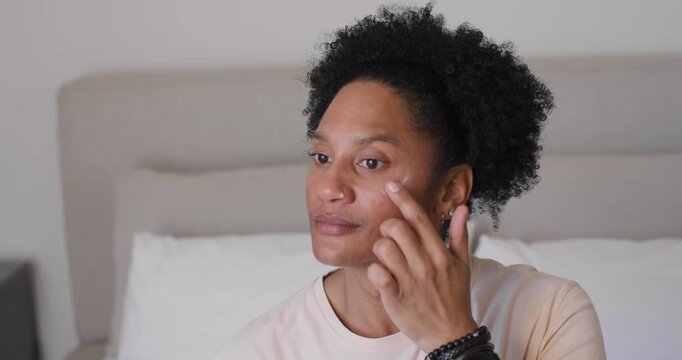Adult African American woman noticing dab under eye, lifting, tapping and blending on bed for care