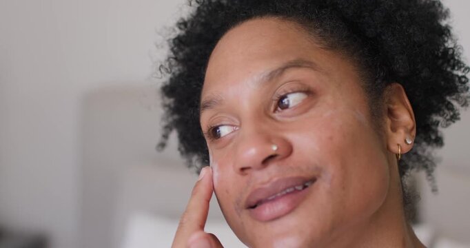 African American woman pressing fingertip with cream, blending upward under-eye in bedroom for care