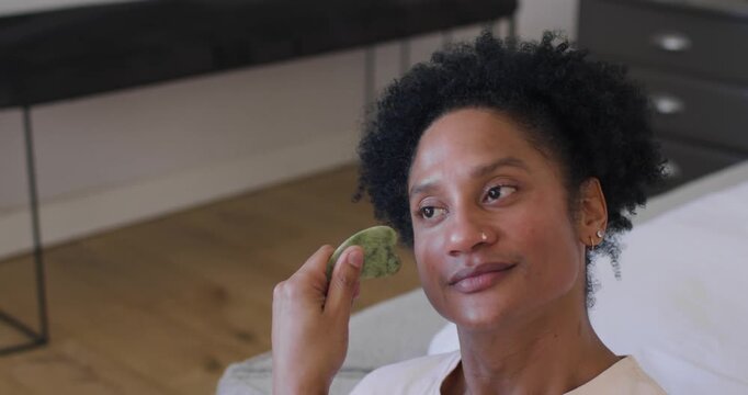 Adult African American woman pressing green stone, sliding toward jawline on sofa for skincare