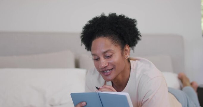 African American man lying on bed holding blue notebook, tapping pen to lips, reading and studying