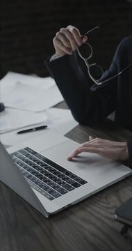 Businesswoman working at laptop in modern office space using touchpad and holding glasses in hand during deep focused work session with documents scattered on desk in cinematic setting.