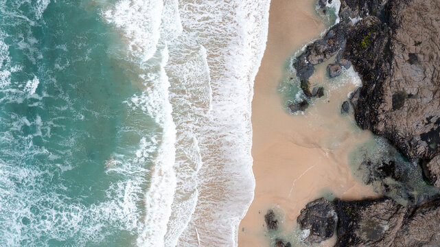 Aerial view of turquoise waves crashing onto the sandy shore and dark coastal rocks at Constantine Bay, Padstow, Cornwall, United Kingdom.