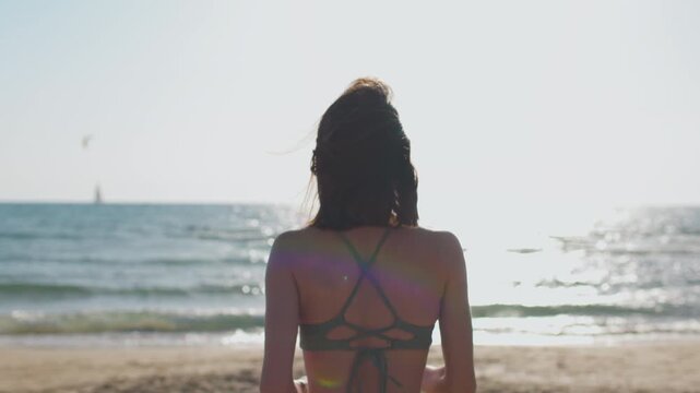 A rear view of a woman running across warm sand towards the sparkling sea, with a brightly lit horizon, gentle waves and a calm summer holiday atmosphere.