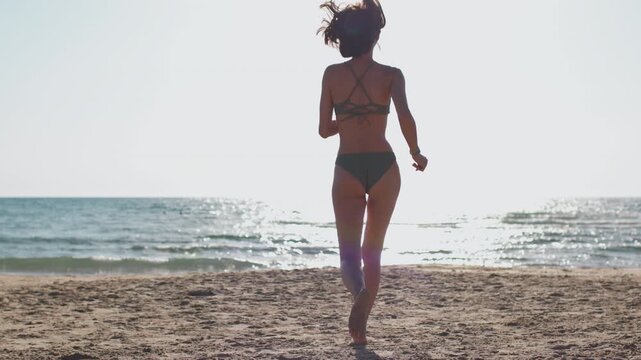 A rear view of a woman running across warm sand towards the sparkling sea, with a brightly lit horizon, gentle waves and a calm summer holiday atmosphere.