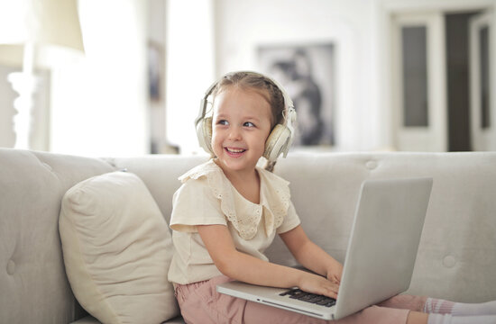 Happy little girl using a laptop with headphones on a sofa
