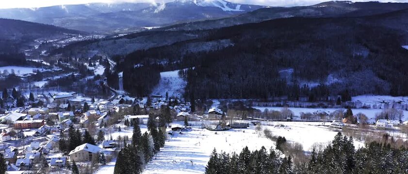 High altitude aerial view showing a picturesque snowy mountain village surrounded by dense winter forests and distant massive ski slopes