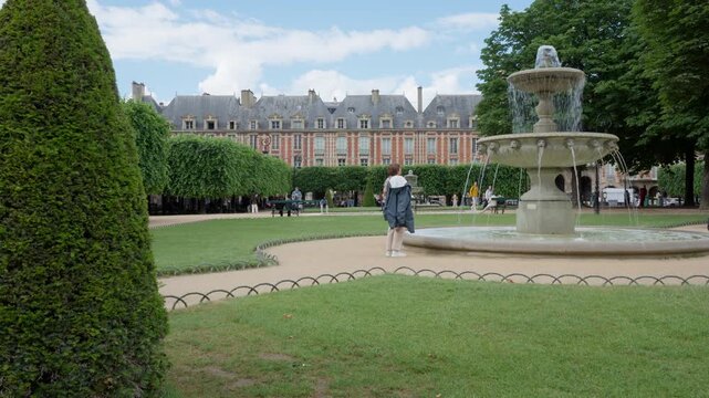 Tourist woman sightseeing at Place des Vosges public square park in Paris France. Historic red brick residential buildings stand behind a flowing multi-tiered stone fountain in summer.