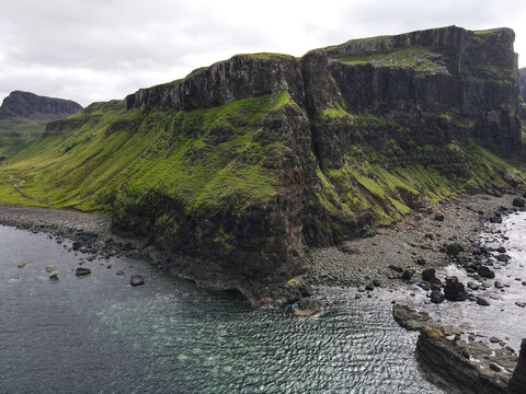 Aerial view of Talisker Bay Beach featuring steep basalt cliffs covered in green grass meeting the rocky shoreline and dark ocean water under an overcast sky Isle of Skye, United Kingdom.