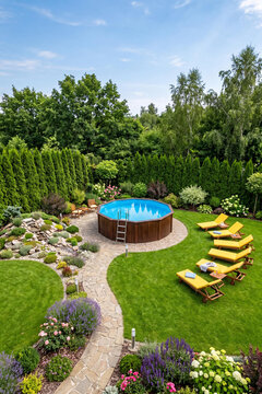 High angle view of a luxury residential backyard featuring a round wooden above-ground pool. Yellow sun loungers on a lush green grass lawn surrounded by beautiful blooming garden flowers.