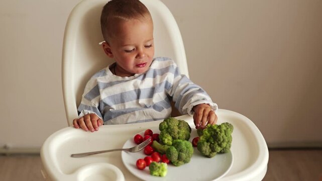 A small child sits in a high chair enjoying broccoli and cherry tomatoes. The setting is a simple kitchen. The child is focused on the food