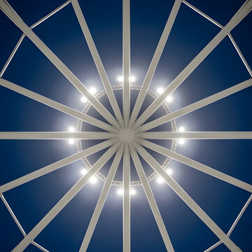 Abstract symmetrical bottom-up view of a circular white steel roof structure with bright lights against a dark blue sky