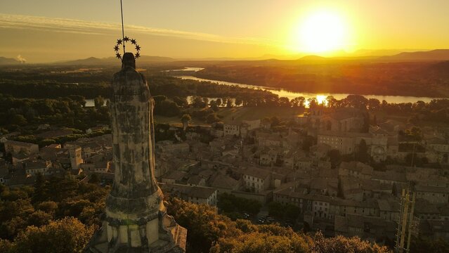 Aerial view of the Virgin Mary statue overlooking the historic town of Viviers, with the Rhone river reflecting the golden sunset light. Viviers, Auvergne-Rhone-Alpes, France.