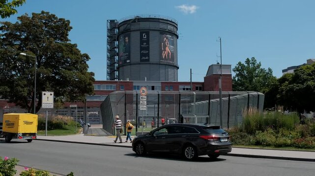 Germany, Wuppertal, June 20 2025. Pedestrians stroll along the sidewalk on a sunny day next to an outdoor soccer cage with a large cylindrical gas holder structure in the background.