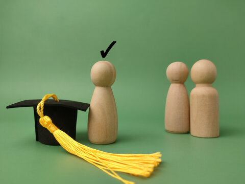 A wooden peg figure stands next to a black graduation cap with a yellow tassel, marked with a check