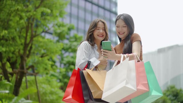 Two happy young Asian women holding colorful shopping bags while having a fun video call on a smartphone in the city. Joyful friends sharing their exciting shopping spree experience outdoors.