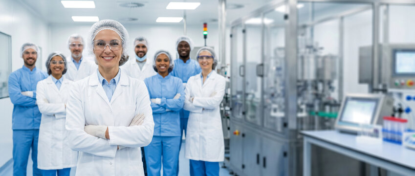 Diverse pharmaceutical manufacturing team standing in modern production facility, led by female pharmaceutical scientist, with chemists, laboratory technicians, and quality control specialists.