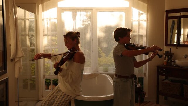 young woman and pre teen boy playing violin in bathroom at sunset