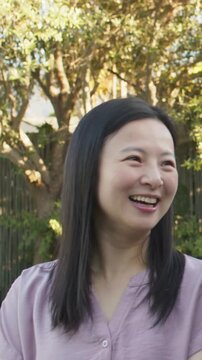 Vertical video: Smiling woman in mauve top looking to camera by fence in backyard, reacting to joke