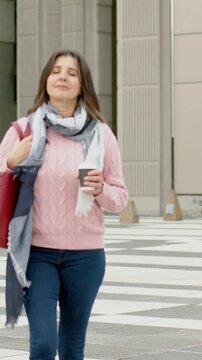 Vertical video: Starting walk woman in pink sweater moving across plaza, holding coffee, red tote