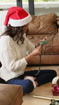 Vertical video: Holding green box and threading ribbon, woman in red Santa hat making gift at home
