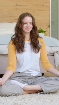 Vertical video: Breathing woman with yellow sleeves, gray pants moving for calm on bedroom rug, bed