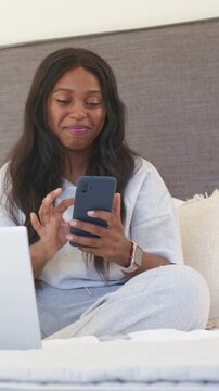 Vertical video: Looking at dark phone, woman in grey sweats scrolling texts on bed with laptop left