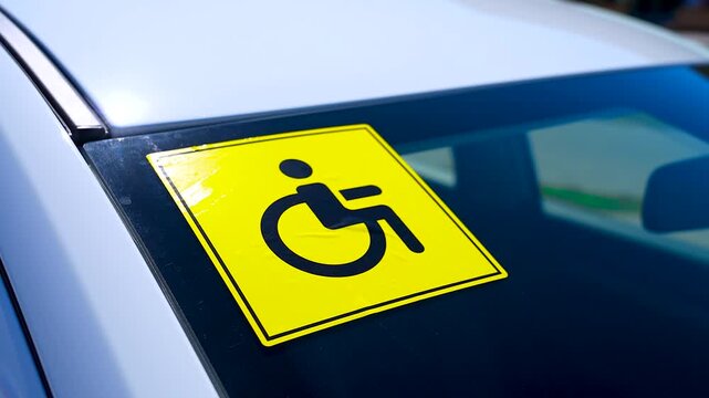 A sticker with the universal wheelchair symbol is glued to the rear window of a white car.