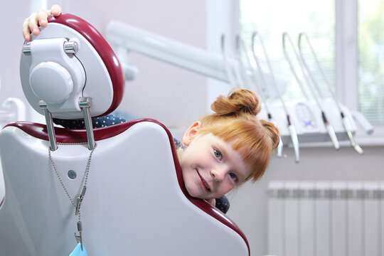 Cheerful girl with red hair is sitting in dental chair. Girl is not afraid of the dentist. Little girl at dentist appointment