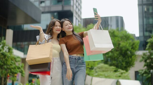 Two happy Asian women posing for a selfie with shopping bags outdoors. Stylish friends enjoying a successful shopping spree at a modern city mall. Lifestyle, fashion, and consumerism concept in urban.