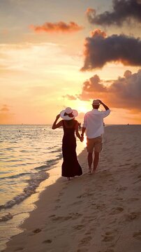 Rear view of a romantic couple on vacations holding hands and walking down a tropical beach during sunset time