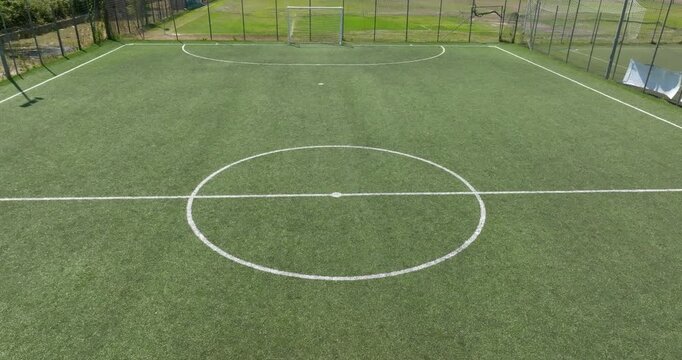 Aerial closeup of a vibrant green synthetic turf soccer field, highlighting the white center circle and midfield line markings, enclosed by a metallic mesh fence. The football pitch is empty. 