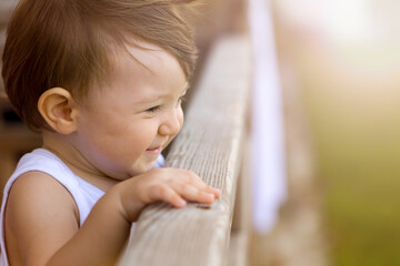 Close up profile portrait of a smiling toddler looking over a wooden railing outdoors, bright sunny day. © Fatih