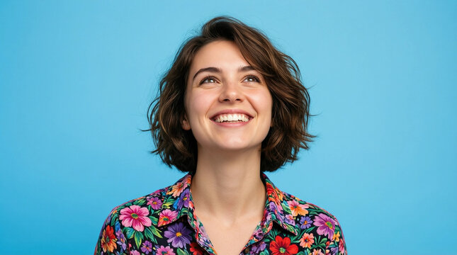 A cheerful woman with short hair smiles upward against a solid blue studio background, wearing a colorful floral shirt.