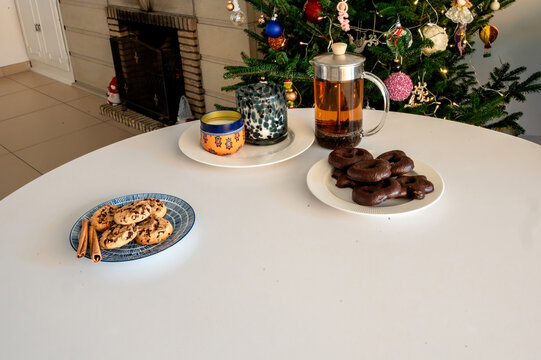 A glass French press of tea, a candle and plates of chocolate gingerbread and chip cookies on a white table before a glowing Christmas tree with a fireplace. A cozy minimal holiday treats and drink