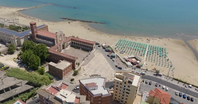 Aerial view of the sanctuary of Our Lady of Grace and Saint Maria Goretti, in Nettuno, Lazio, Italy. It stands at the end of the seafront, along the coast, next to a sandy beach lined with umbrellas.