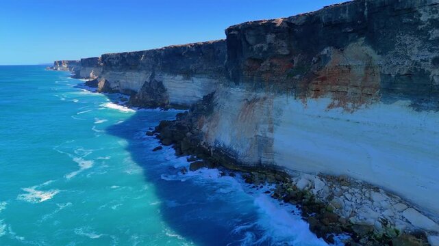 Aerial drone shot approaching the sheer Bunda Cliffs from above the ocean with blue sky, blue water, and white waves crashing below