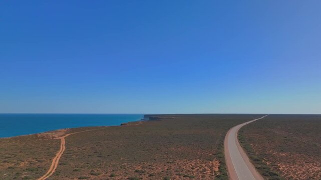 Aerial drone shot rising backward over the Australian outback with a lone road stretching to the horizon and distant Bunda Cliffs along the coastline