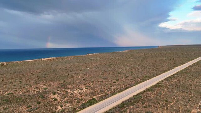 Aerial view following a diagonal road over open wilderness with the Bunda Cliffs, ocean waves, and a bright rainbow on the horizon