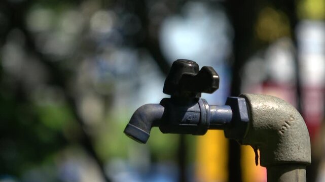A hand attempts to open a blue plastic spigot attached to a metal pipe on the park, but no water discharges.