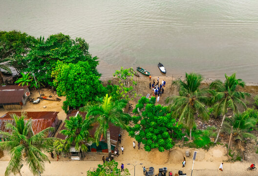Aerial view of people gathered near small boats on the riverbank surrounded by lush palm trees and simple structures in Minima, Rivers, Nigeria.