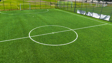 Aerial closeup of a vibrant green synthetic turf soccer field, highlighting the white center circle and midfield line markings, enclosed by a metallic mesh fence. The football pitch is empty.  © Stefano Tammaro